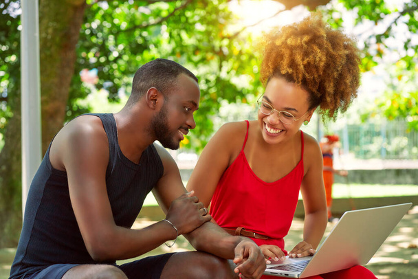 dois jovens adultos negros olhando e usando um laptop e rindo em um parque ao ar livre - Foto, Imagem