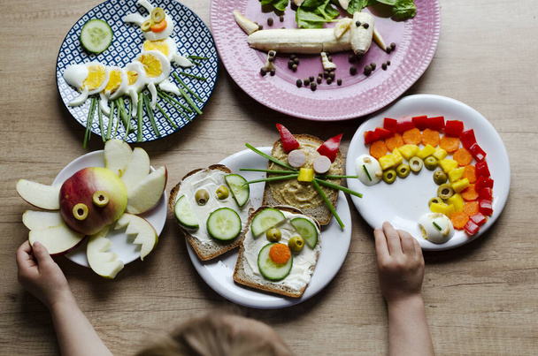 Fun with food with little child. Colorful table with different vegetables and healthy kids food. Hands of child with plates of creative breakfast. - Photo, image
