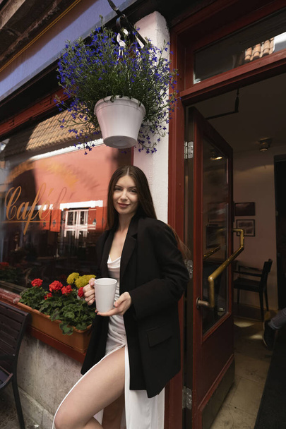 Beyaz elbiseli ve siyah ceketli güzel bir esmer, Avrupa 'nın dar bir sokağındaki açık kafede kahve içiyor. - Fotoğraf, Görsel