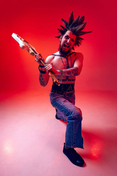 Crazy punk. Emotional young man, rock musician posing aggressively with electric guitar against red studio background in neon light. Concept of music, lifestyle, subculture, art, youth, human emotions - Φωτογραφία, εικόνα