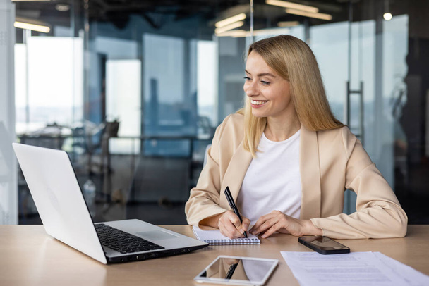 Young smiling and successful businesswoman, manager works in the office with a laptop online, talks on a video call, listens and watches business training, makes notes in a notebook. - Foto, Bild