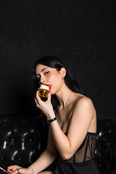 portrait Elegantly dressed young beautiful girl with black hair and dress celebrating 18th birthday. sitting on black leather couch eating little cake. black background. - Photo, Image