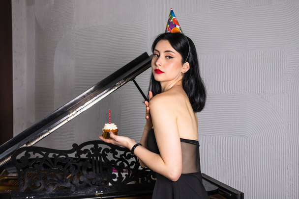 Elegánsan öltözött fiatal gyönyörű lány fekete haj és ruha ünnepli 18. születésnapját, pózol zongora ellen - Fotó, kép