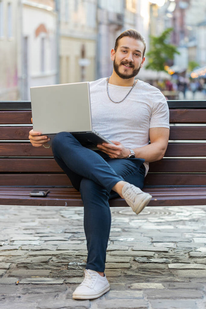 Happy smiling man opens laptop sitting on bench in urban city street outdoors. Middle eastern guy works on notebook, sends messages, makes online purchases, watching movies. Town lifestyles. Vertical - Zdjęcie, obraz