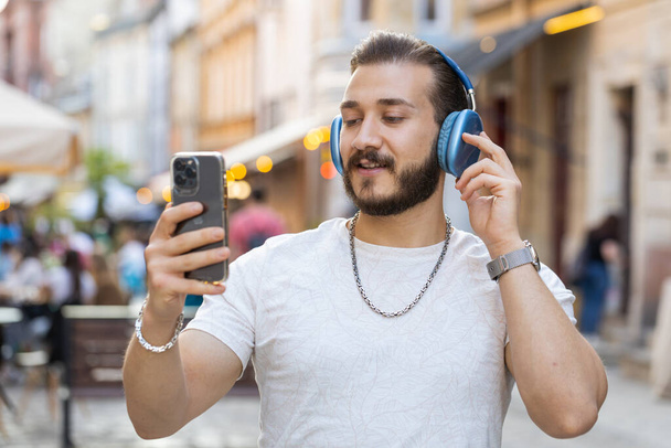 Happy relaxed overjoyed bearded young man in wireless headphones choosing, listening favorite energetic disco rock n roll music dancing outdoors. Guy walking in urban city sunshine street background - Photo, Image