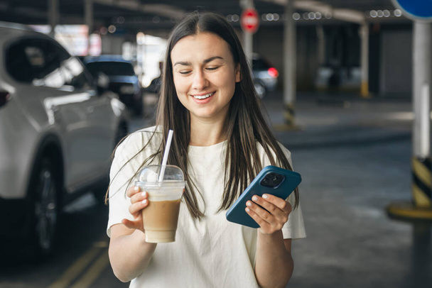 Cheerful young woman with a cup of coffee and a smartphone in her hands in a car parking. - Foto, imagen