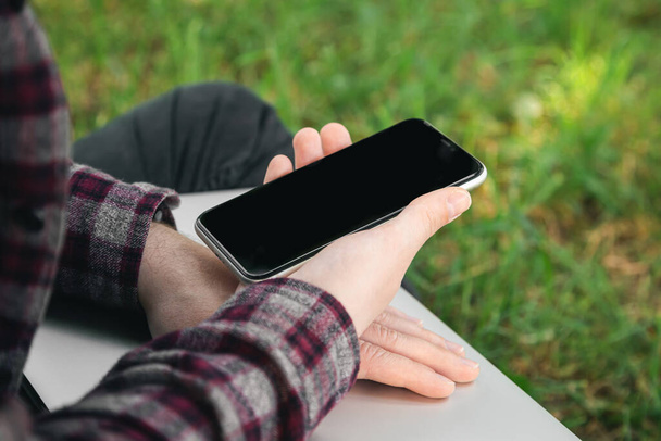 Een man zit op het gras met een laptop en een smartphone in zijn handen, met behulp van gadgets buiten, close-up. - Foto, afbeelding