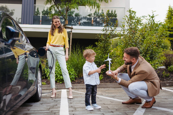 Happy family standing by beside their electric car and charging it on the street. Electric vehicle with charger in charging port. Little boy with dad playing with model of wind turbine. - Zdjęcie, obraz