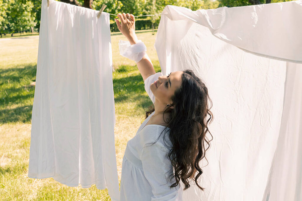 Urocza dziewczyna, gospodyni domowa w jasnobiałej sukience i opasce, wisi czyste pranie do wyschnięcia na zewnątrz w ciepły letni dzień. - Zdjęcie, obraz