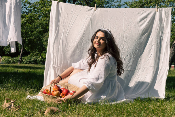 A lovely girl, a homemaker in a light white dress and headband, hangs clean laundry to dry outside on a warm summer day, arranged a picnic, sunbathes, and enjoys eating fruits. - Photo, Image