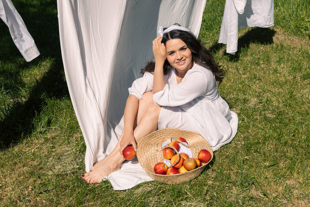 Hoş bir kız, açık beyaz elbiseli ve bantlı bir ev hanımı, sıcak bir yaz gününde dışarıda kurutmak için temiz çamaşırlarını asar, bir piknik düzenler, güneşlenir ve meyve yemekten zevk alır.. - Fotoğraf, Görsel