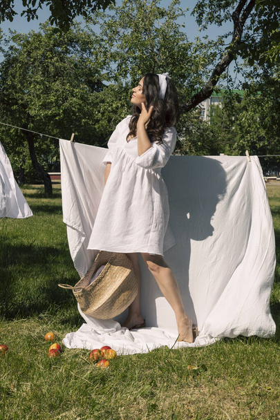 A lovely girl, a homemaker in a light white dress and headband, hangs clean laundry to dry outside on a warm summer day, arranged a picnic, sunbathes, and enjoys eating fruits. - Photo, Image
