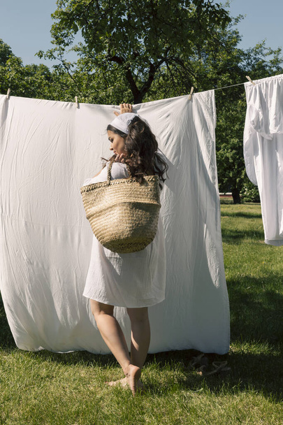 A lovely girl, a homemaker in a light white dress and headband, hangs clean laundry to dry outside on a warm summer day. - Фото, изображение