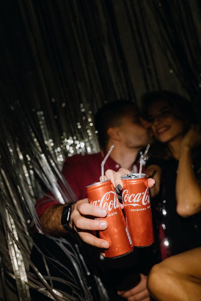 CHERNIGIV, UKRAINE - NOVEMBER 21, 2022: A young couple sits on the floor and kisses while holding Coca-Cola against a curtain of gray rain. High quality photo - Fotografie, Obrázek