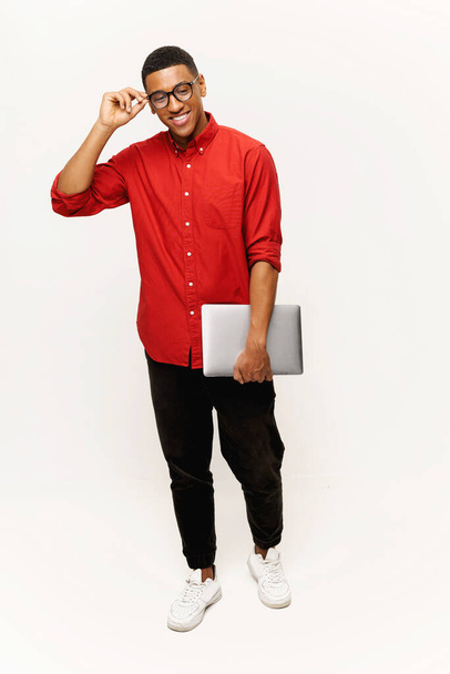 Vertikale Aufnahme eines optimistischen gutaussehenden afroamerikanischen Unternehmers in lässigem rotem Hemd und stilvoller Brille, der mit Laptop auf weißem Hintergrund steht, ausländischer männlicher Student - Foto, Bild
