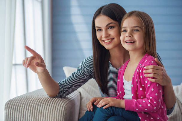 Beautiful young mother and her cute little daughter are smiling, pointing and looking away, sitting on sofa at home - Фото, изображение