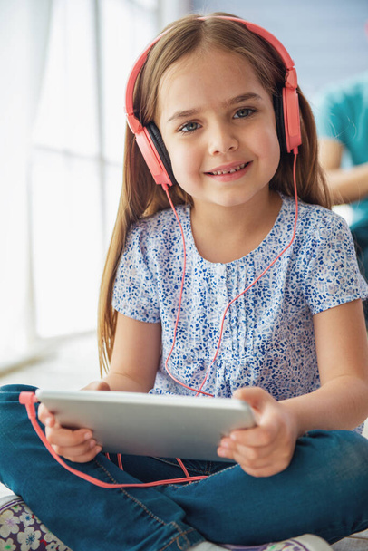 Portrait of cute little girl in headphones listening to music using a tablet, looking at camera and smiling while sitting on the floor - Photo, Image
