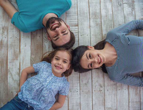 Top view of beautiful young parents and their cute little daughter looking at camera and smiling, lying on wooden floor - Photo, Image