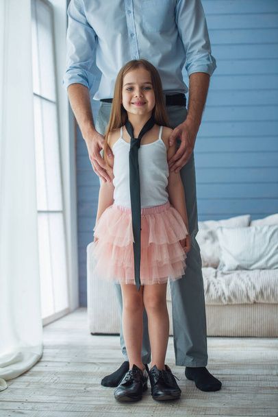 Das Bild zeigt einen jungen Vater in klassischer Kleidung und seine süße kleine Tochter, die im Raum steht. Mädchen in väterlicher Krawatte blickt in die Kamera und lächelt - Foto, Bild