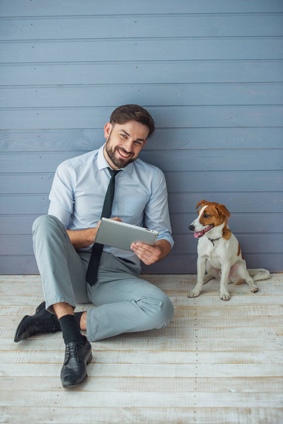Knappe jonge zakenman is met behulp van een tablet, camera kijken en glimlachen terwijl zittend op de vloer met zijn hond - Foto, afbeelding