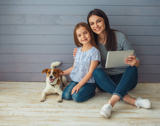 Милая маленькая дочь и ее красивая молодая мама смотрят в камеру и улыбаются, позируя со своей собакой. Мама использует планшет
 - Фото, изображение