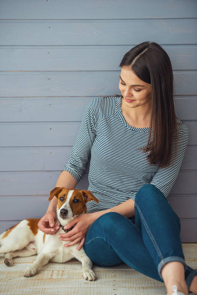 Beautiful young woman is smiling while sitting with her dog on the floor - Photo, Image