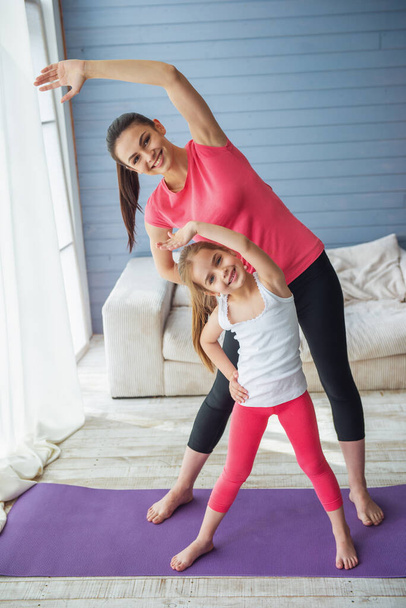Beautiful young mother and her cute little daughter are looking at camera and smiling while stretching on yoga mat at home - Photo, Image