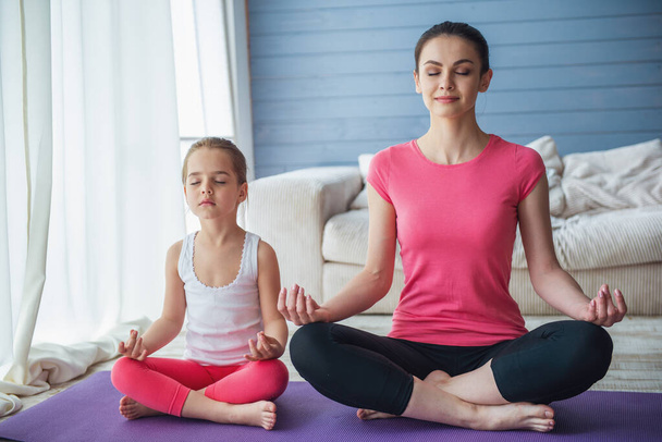Die schöne junge Mutter und ihre süße kleine Tochter lächeln und meditieren, während sie zu Hause in Lotuspose auf der Yogamatte sitzen - Foto, Bild