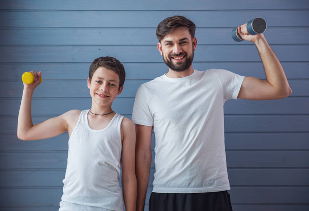 Guapo joven padre y su hijo adolescente están mirando a la cámara y sonriendo mientras se ejercita con pesas
 - Foto, imagen