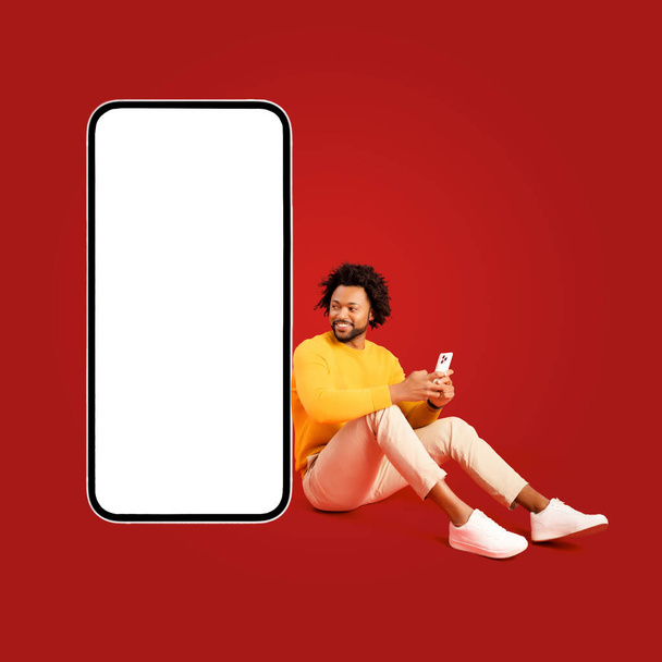 Komea Afrikkalainen-amerikkalainen kihara 30s mies käyttää älypuhelinta istuu lähellä valtava matkapuhelin tyhjä näyttö eristetty punainen, karismaattinen kaveri suosittelee ja mainonta mobiilisovellus, kopioi tilaa - Valokuva, kuva