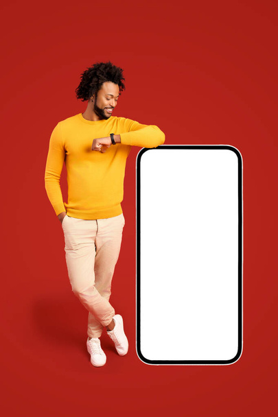 Best deal, promotion. Smiling positive african-american man leaning on huge smartphone with empty blank screen, looking at it, freelancer guy recommending new app or website, mockup - Foto, afbeelding