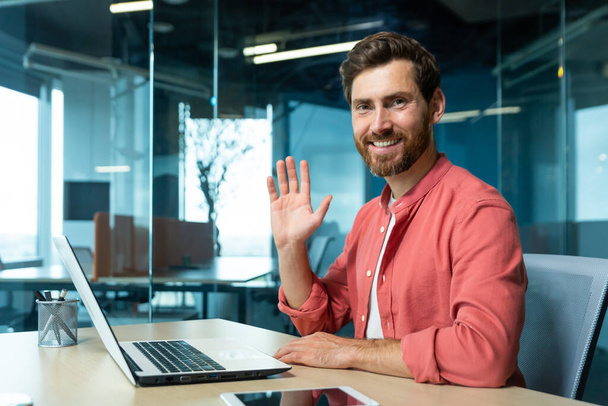 Portrait of successful financier businessman inside office at workplace, man smiling and looking at camera waving hand gesture of greeting, small business owner working using laptop - Photo, image