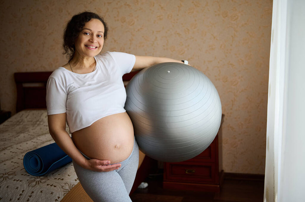 Happy multi ethnic pregnant woman, expectant gravid mother with big belly in late pregnancy, holding a fitball, touching her tummy, smiling looking at camera, enjoy morning prenatal fitness workout - 写真・画像