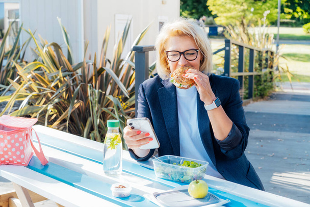 Middle-aged business woman eating a wholemeal burger with salad and using a phone sitting at picnic table in the park while having a break. Healthy lunch box for weight loss. Balanced diet. - Valokuva, kuva