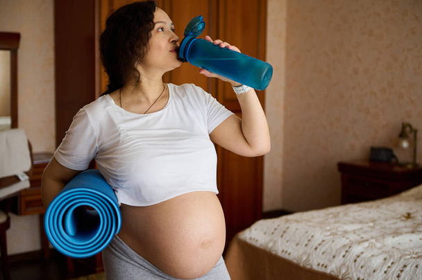 Multi ethnic active sporty pregnant woman in crop white t-shirt with naked big belly in late pregnancy, expectant mother drinking water from sports bottle after prenatal fitness and stretching workout - Photo, Image