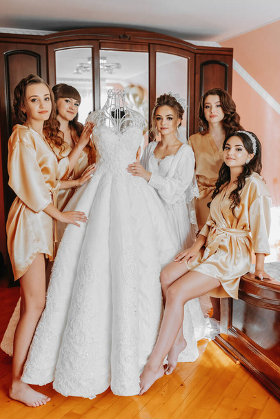 Young bridesmaids in silk robes in a hotel room. Beautiful women celebrate a bachelorette party, next to a luxurious dress of the bride in themed pajamas. - Foto, imagen