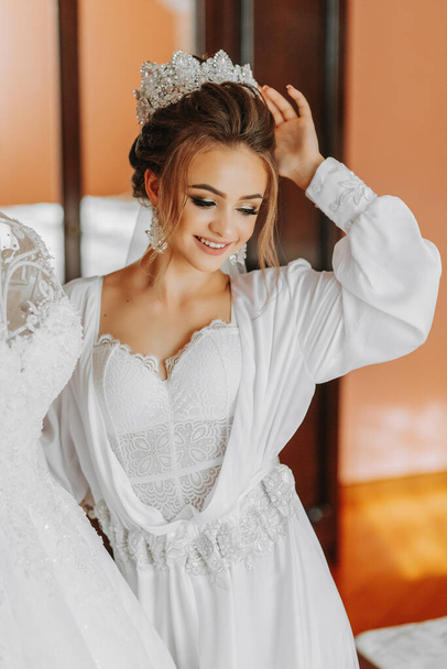 A happy young bride with a chic royal hairstyle and delicate makeup near her dress. Royal tiara, white petticoat, portrait photo - Фото, изображение