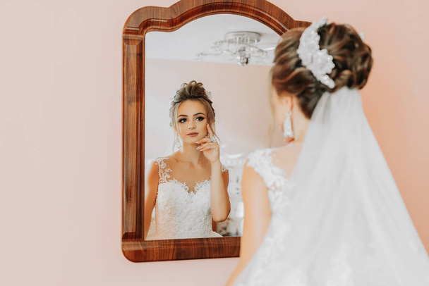 A menyasszony egy gyönyörű ruhában belenéz a tükörbe, az utolsó előkészületek. Esküvő. esküvői csokor A menyasszony reggele. Egy boldog menyasszony vár a vőlegényre.. - Fotó, kép