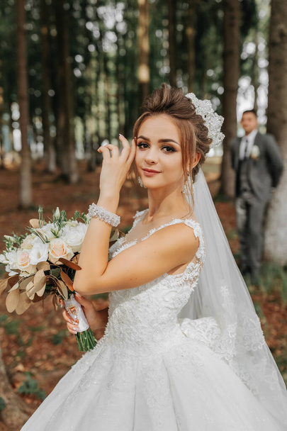 A beautiful young woman in a wedding dress between tall trees in the forest with a royal hairstyle and a chic tiara with a bouquet of flowers in her hands, a wedding in golden color - Fotoğraf, Görsel