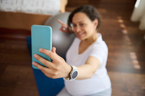 Primer plano de teléfono móvil inteligente en las manos de la mujer embarazada, embarazada gravid madre tomando selfie sentado en una alfombra de ejercicio después del entrenamiento de acondicionamiento físico prenatal en casa. Estilo de vida activo embarazo saludable - Foto, imagen