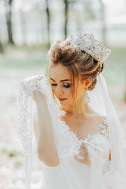 Beautiful young woman in a wedding dress in the forest with a royal hairstyle and a chic tiara holds her veil in her hands, wedding in gold color - Photo, Image