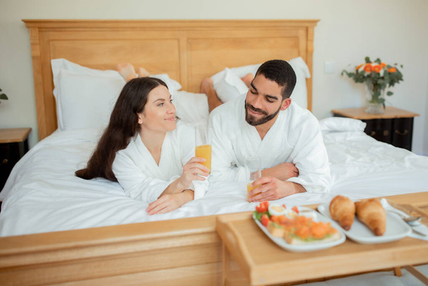 Hotel Room Service. Relaxed Couple Enjoying Romantic Breakfast In Bed, Drinking Fresh Orange Juice Lying Wearing Bathrobes In Bedroom Interior, Near Tray With Delicious Food. Honeymoon Vacation - Foto, immagini