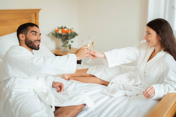 Romantic Honeymoon. Newlywed Couple Sharing Intimate Moment, Clinking Champagne Glasses, Enjoying Sparkling Wine Together In Hotel Bedroom Interior. Vacation Leisure And Relax - Photo, Image