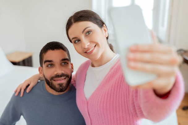 Social Media Aanwezigheid. Vrolijke Bloggers paar maken Selfie Holding Smartphone In de hand, poseren Omarmen In Modern Slaapkamer Interieur. Selectieve focus op jonge echtgenoten. Gadgets en Online Plezier - Foto, afbeelding