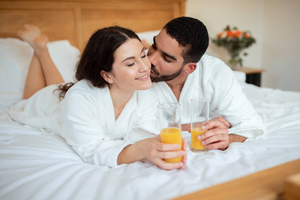 Echtgenoot kussen terwijl vrouw ze nippen sinaasappelsap, met een frisse ochtend drinken en romantisch ontbijt in bed in Luxury Hotel. Echtgenoten Ontspannen Genieten van geweldige roomservice op vakantie Accommodatie - Foto, afbeelding