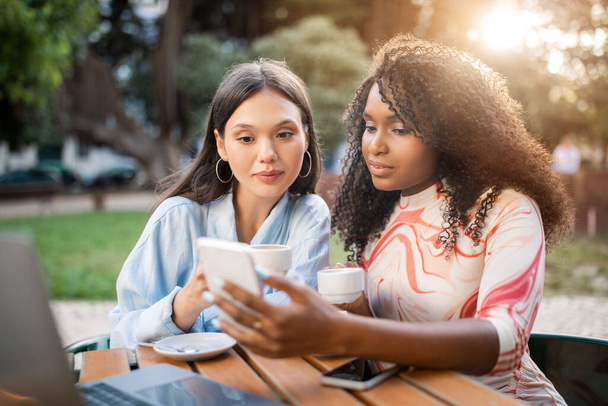 Kaksi nuorta naista rentoutuu älypuhelimen kanssa ulkona kahvilassa, onnelliset monikansalliset naisystävät juovat kahvia ja tarkistavat uuden verkkokaupan, käyttävät matkapuhelinta lepäämällä pöydässä ulkona, lähikuva - Valokuva, kuva
