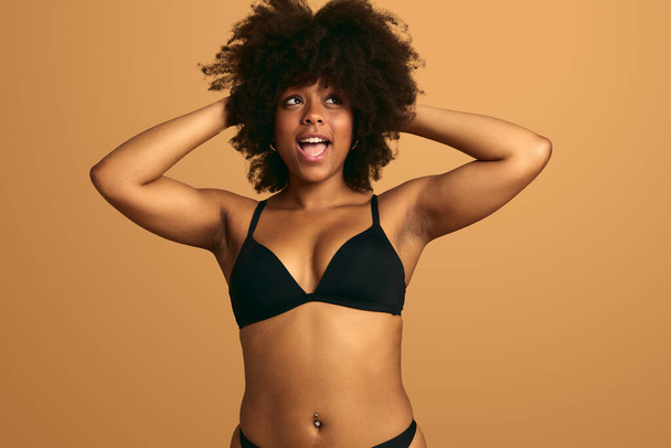 Happy curvy African American woman with Afro hair in black lingerie standing on beige background and touching head while looking away - Фото, изображение