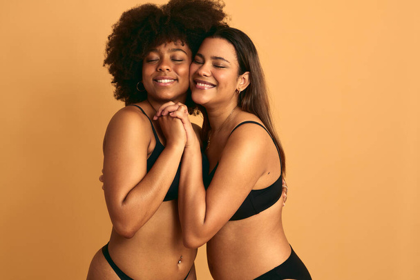 Cheerful young multiracial female models in black underwear embracing each other and smiling with closed eyes, while standing close and holding hands against beige background together in studio - Foto, Bild