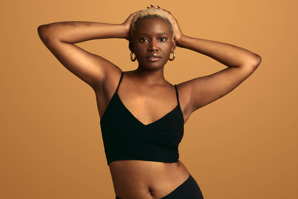 Young African American female model with short blond hair wearing black underwear touching head while looking at camera against orange background - Foto, afbeelding