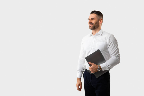 Cheerful handsome middle aged businessman with modern pc laptop in his hand walking towards copy space over light studio background. Online business offer, deal, discount, sale - Valokuva, kuva